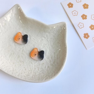 May include: Two small, two-hole buttons shaped like cat heads. Each button features a gradient color pattern transitioning from orange on one side to black on the other. The buttons are smooth and have a glossy finish, with two small holes for sewing.