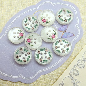 May include: Nine white, round buttons with six holes each, decorated with pink roses and green leaves. The buttons are arranged on a lavender card with the words "SMILE EVERYDAY" printed on it. A butterfly sticker is also visible.