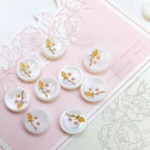May include: A collection of white, pearlescent buttons with a floral design. Each button features a gold-colored embroidered rose on a stem. The buttons have two holes for sewing and are arranged on a pink floral-themed background.