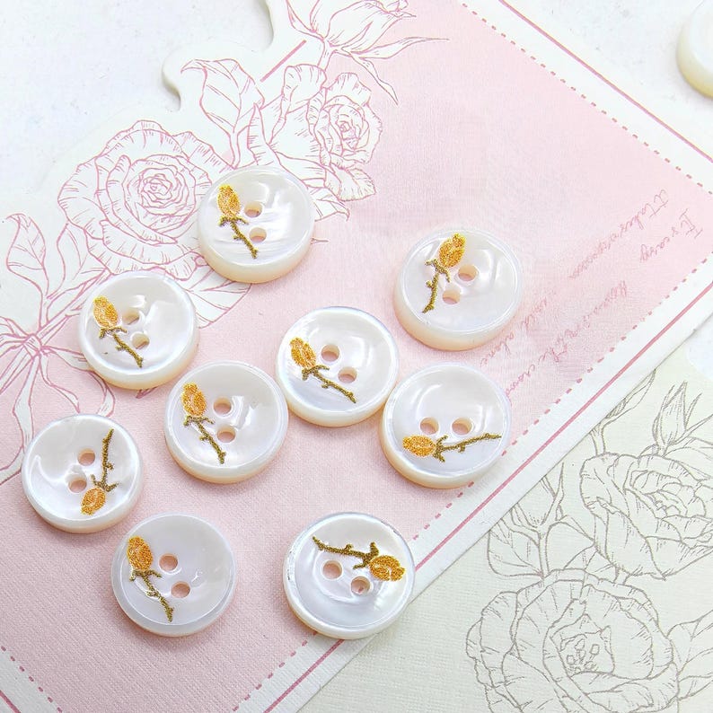 May include: A collection of white, pearlescent buttons with a floral design. Each button features a gold-colored embroidered rose on a stem. The buttons have two holes for sewing and are arranged on a pink floral-themed background.