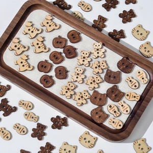 May include: A collection of wooden buttons in diverse shapes, such as teddy bears, cats, apples, and fish, displayed on a wooden tray. The buttons are in light and dark brown hues, featuring buttonholes for sewing.