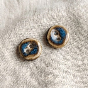 May include: Two round buttons featuring a glossy finish with a central blue circular pattern surrounded by a tan and brown gradient border. Each button has two holes in the centre for attachment, showcasing a smooth, reflective surface texture.