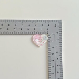 May include: A heart-shaped resin charm with a pink and white gradient, featuring cartoon dog and bear illustrations. The charm is approximately 1 inch in width and is filled with small, decorative elements. A ruler is placed next to the charm.