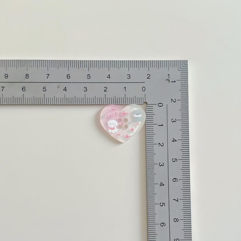 May include: A heart-shaped resin charm with a pink and white gradient, featuring cartoon dog and bear illustrations. The charm is approximately 1 inch in width and is filled with small, decorative elements. A ruler is placed next to the charm.