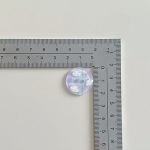 May include: A round button with a pastel purple and blue background. The button has a design of white sheep and pink bows. The button measures approximately 0.75 inches in diameter and has two holes for sewing.