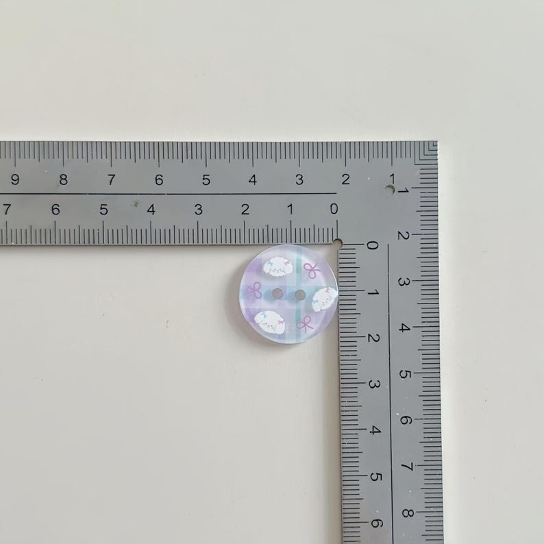 May include: A round button with a pastel purple and blue background. The button has a design of white sheep and pink bows. The button measures approximately 0.75 inches in diameter and has two holes for sewing.