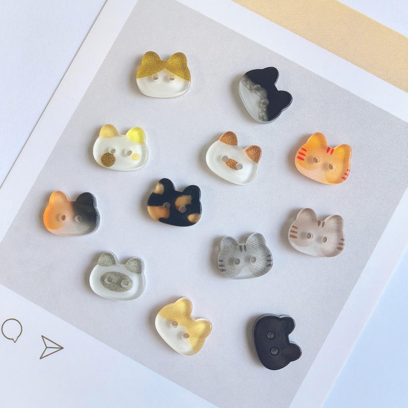May include: A collection of twelve small, cat-shaped buttons made of translucent and opaque resin. Each button features distinct color patterns including solid black, white, orange, grey, and tortoiseshell designs, with two small holes for sewing.