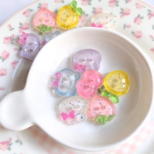 May include: A collection of small, translucent, two-hole buttons shaped like fruits and animals, featuring glitter accents and small colored bows. The buttons are in shades of pink, yellow, blue, and purple, and are scattered inside and around a white bowl.