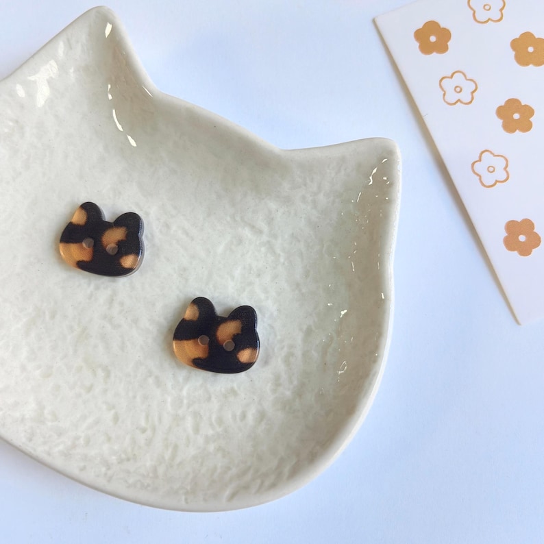 May include: Two small, flat buttons shaped like cat heads with a mottled tortoiseshell pattern in shades of black, brown, and tan. Each button features two small holes in the center for attachment, showcasing a smooth, glossy finish on the surface.