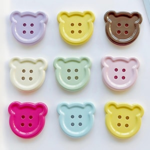 May include: Nine bear-shaped buttons in pastel colours. Arranged in a three-by-three grid, each button has four small holes for sewing. Colours include lavender, yellow, brown, cream, green, pink, fuchsia, blue, and light yellow. Ideal for craft projects.