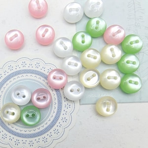 May include: An assortment of iridescent, oval buttons in pastel shades of pink, white, green, and yellow. Each button features two sewing holes. The buttons are arranged on a decorative paper doily.