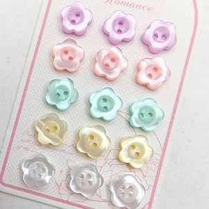 May include: A collection of flower-shaped buttons in various pastel colors, including pink, purple, green, and yellow. Each button has a pearlescent sheen and a two-hole design for sewing. The buttons are arranged on a decorative card.