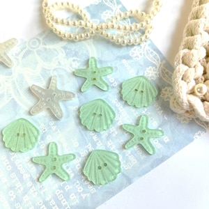 May include: A selection of sea-themed buttons shaped like starfish and seashells. The buttons are a translucent, pale green colour with two holes for sewing. A string of pearls and a white rope-like object are also in the image.