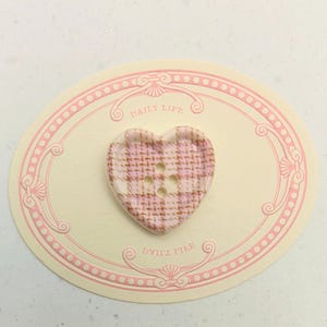 Op de afbeelding: Een hartvormige knoop met een roze en witte ruitpatroon. De knoop heeft vier gaatjes en ligt op een cr&egrave;mekleurige ovale kaart met roze sierranden en de woorden "DAILY LIFE" en "DVITA TILE".