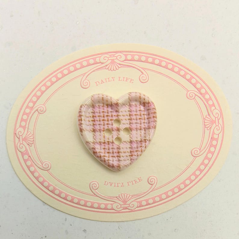 Op de afbeelding: Een hartvormige knoop met een roze en witte ruitpatroon. De knoop heeft vier gaatjes en ligt op een cr&egrave;mekleurige ovale kaart met roze sierranden en de woorden "DAILY LIFE" en "DVITA TILE".