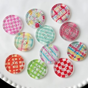 May include: A collection of colorful, transparent buttons with various patterns. Designs include plaid, stripes, and whimsical illustrations in shades of pink, red, green, blue, and yellow. Each button has four holes for sewing.