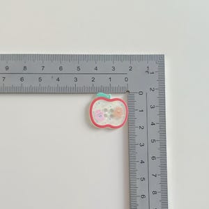 May include: An apple-shaped button with a red outline, green stem, and clear center. Inside are two small animal figures and three small, round, gray buttons. The button measures approximately 1 inch in length.