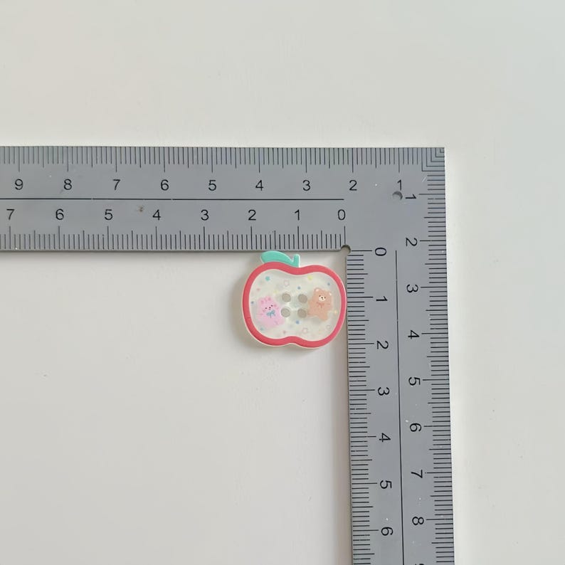 May include: An apple-shaped button with a red outline, green stem, and clear center. Inside are two small animal figures and three small, round, gray buttons. The button measures approximately 1 inch in length.