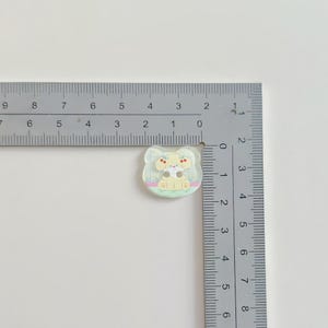 May include: A small, clear, decorative button with a cartoon dog design. The dog is light yellow with brown spots and a yellow bow. The button is approximately 1 inch in size and sits on a white surface next to a ruler.