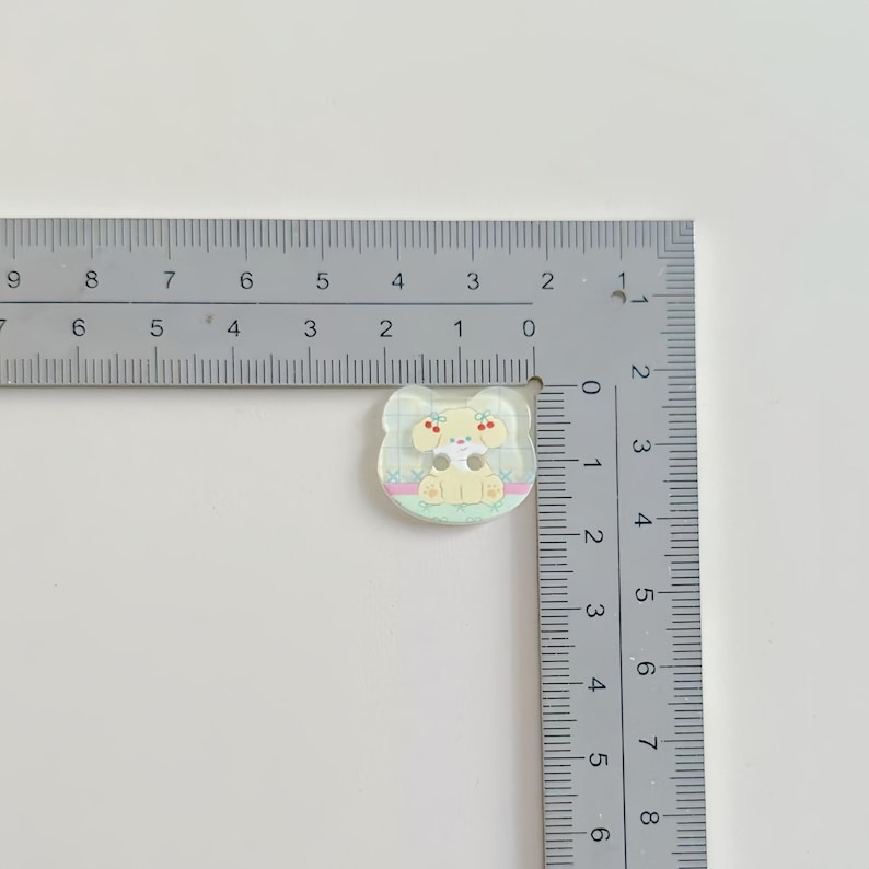 May include: A small, clear, decorative button with a cartoon dog design. The dog is light yellow with brown spots and a yellow bow. The button is approximately 1 inch in size and sits on a white surface next to a ruler.
