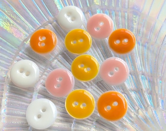 May include: A collection of colorful, glossy buttons in various shades of white, pink, orange, and yellow. Each button has two holes and a rounded shape. The buttons are arranged on a reflective, iridescent surface.
