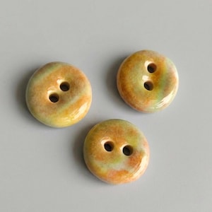 May include: Three round buttons with two holes each. The buttons have a marbled appearance with shades of yellow, orange, and green. The buttons are glossy and appear to be made of ceramic or a similar material. The buttons are for sewing or crafts.