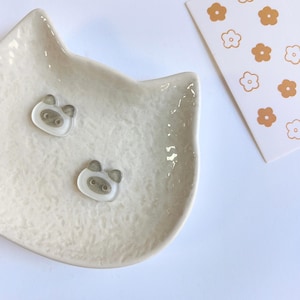 May include: A cream-colored ceramic dish shaped like a cat head holds two small, translucent, light gray pig-shaped charms. Beside the dish is a white rectangular card featuring a repeating pattern of small, solid, and outlined orange flowers.