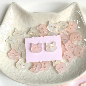 May include: A collection of cat-shaped buttons and a pair of cat-face earrings. The buttons and earrings are in shades of pink and white, with a pearlescent sheen. They are displayed on a cat-shaped dish.