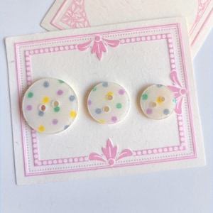 May include: Three round, off-white buttons with colourful polka dots in yellow, green, purple, and blue. The buttons are arranged on a decorative card with a pink border and floral accents. The buttons vary in size.