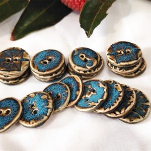 May include: A collection of handmade ceramic buttons. The buttons are round with two holes, featuring a glossy turquoise glaze and a brown rim. The buttons are stacked in small piles and arranged on a white surface.