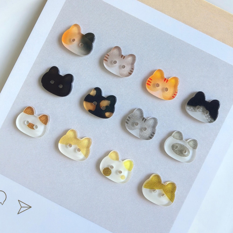 May include: A collection of twelve cat-shaped buttons in various colors and patterns, including solid black, tortoiseshell, orange tabby, grey tabby, and white with gold or silver glitter accents. Each button features two holes for sewing attachment.