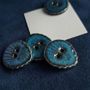 May include: Close-up of several handmade, ceramic buttons. The buttons are a vibrant teal color with a textured surface and a dark gray rim. Each button has two holes for sewing. The buttons are arranged on a dark blue fabric.