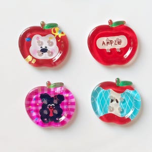 Può includere: Quattro ciondoli in resina a forma di mela in rosso, rosa e blu. Ogni ciondolo presenta un design diverso: un orso bianco, la parola "APPLE", un orso nero e una mela morsa. I ciondoli hanno gambi verdi.