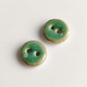 May include: Two round ceramic buttons featuring a glossy green centre glaze that transitions into a light beige or tan rim. Each button has two central holes for attachment, with a smooth, slightly irregular surface texture and visible white speckles.