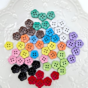 Op de afbeelding: Een verzameling bloemvormige knopen in verschillende kleuren, waaronder groen, bruin, oranje, blauw, wit, paars, geel, roze, zwart en rood. Elke knoop heeft vier gaten om op te naaien. De knopen zijn gerangschikt op een wit, decoratief oppervlak.