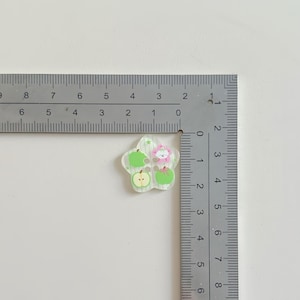 May include: A clear, flower-shaped embellishment with green apples and a pink flower design. The embellishment is approximately 1 inch in diameter and is placed next to a ruler for scale.