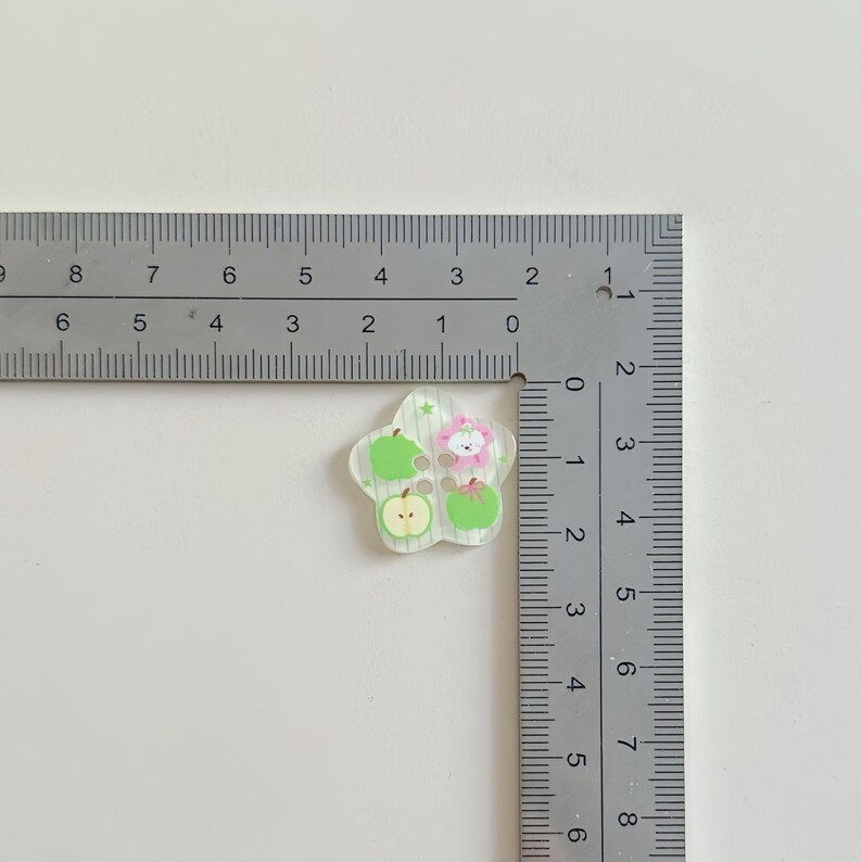 May include: A clear, flower-shaped embellishment with green apples and a pink flower design. The embellishment is approximately 1 inch in diameter and is placed next to a ruler for scale.