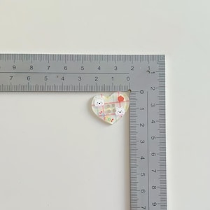 May include: A heart-shaped acrylic charm featuring a pastel plaid pattern, two white dog illustrations, a small red apple, and the word "sweet" in colorful letters. A metal ruler shows the charm measures approximately 1 inch in width.