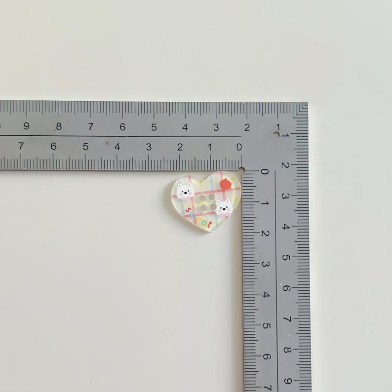 May include: A heart-shaped acrylic charm featuring a pastel plaid pattern, two white dog illustrations, a small red apple, and the word "sweet" in colorful letters. A metal ruler shows the charm measures approximately 1 inch in width.