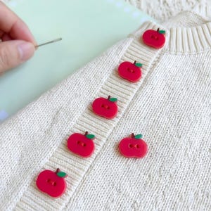 May include: A close-up of a cream-coloured knitted cardigan with red apple-shaped buttons. The buttons have green leaf details. A needle is held near the fabric, suggesting sewing or repair.