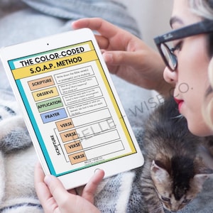 Op de afbeelding: Een witte tablet toont "THE COLOR-CODED S.O.A.P. METHOD" met secties voor Schrift, Observeren, Toepassing en Gebed. De tablet wordt vastgehouden door een persoon met een kitten in de buurt. Het scherm van de tablet heeft een kleurrijk ontwerp.
