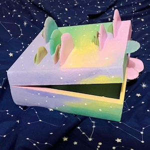 May include: A pastel-colored, open wooden box with a landscape design. The box is painted with a gradient of colors, including purple, green, yellow, and pink. The top of the box features small, three-dimensional mountain-like shapes in various colors.