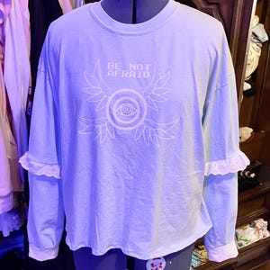 Puede incluir: Una camisa de manga larga azul claro con un diseño blanco de ojo y alas. La camisa tiene un ribete de encaje blanco en las mangas y dice "BE NOT AFRAID" en texto blanco.