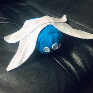 Op de afbeelding: Een handgemaakt blauw pluche speelgoed met witte vleugels en drie witte knopen. Het speelgoed is gemaakt van zachte stof en heeft een grillig ontwerp, dat lijkt op een gestileerd insect of wezen.