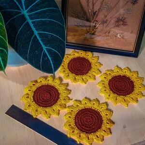 Può includere: Quattro sottobicchieri a forma di girasole all'uncinetto fatti a mano con petali gialli e un centro marrone sono esposti su una superficie di legno. Un righello è parzialmente visibile. Una grande foglia verde e una stampa botanica incorniciata sono sullo sfondo.