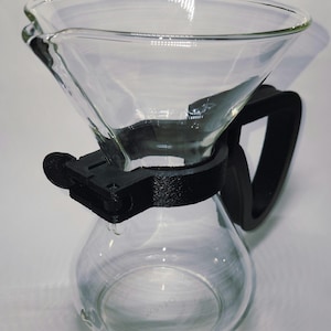 May include: A clear glass pour-over coffee maker with a black plastic handle and support ring. The carafe has a rounded bottom and a conical top for brewing coffee. The design is simple and functional, suitable for home or cafe use.
