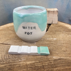 May include: A handmade ceramic water pot with a turquoise glaze dripping down the sides. The words "WATER POT" are printed on the front. A small, white ceramic tray with a turquoise accent sits in front of the pot. The items are on a wooden surface.