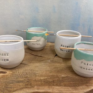 May include: Four handmade ceramic paint water cups in white and mint green, each with a wooden paintbrush and the words "PAINT WATER" embossed on the side. The cups are on a wooden surface.