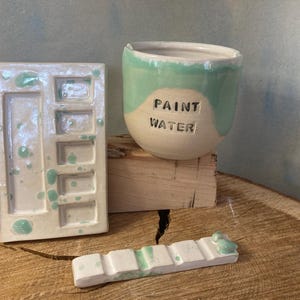 May include: A ceramic paint water cup with the text PAINT WATER, a rectangular paint palette with multiple wells, and a long brush rest. All items are off-white with light green speckled glaze accents and a smooth, glossy finish.