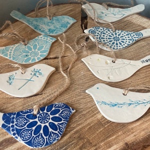 May include: A collection of seven flat, bird-shaped ceramic ornaments with jute twine loops for hanging. Each piece features unique blue and white patterns, including floral motifs, geometric mandalas, and botanical prints, with one bird clearly stamped with the word Mum.